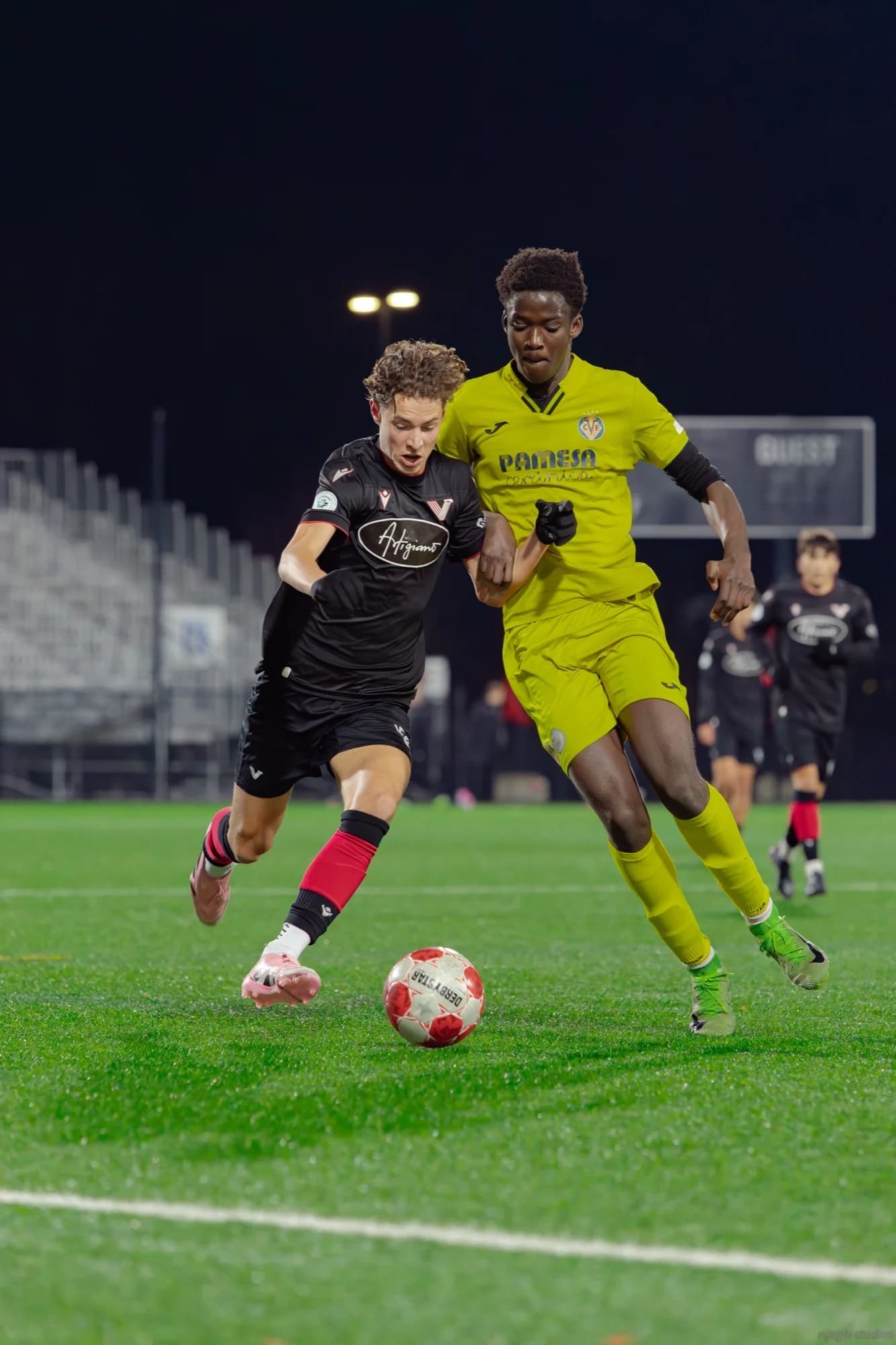 Night soccer match with two players battling for possession under floodlights - Njagih Studios Vancouver
