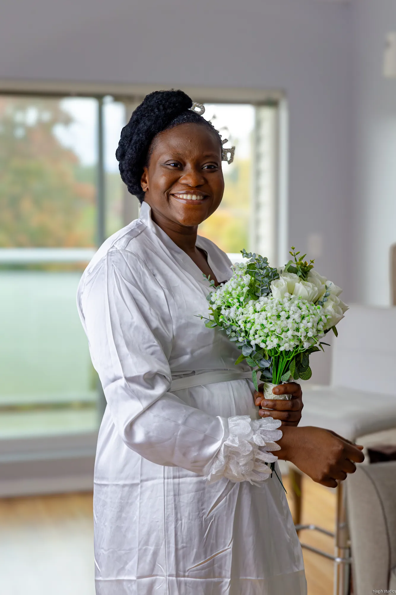 Bride smiling in robe holding bouquet of white roses during bridal preparation - Njagih Studios wedding photography